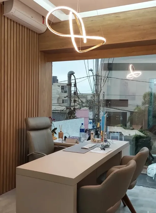Escritório com mesa de atendimento em tom claro, ripado amadeirado e luminária pendente em fita de LED, junto a uma vitrine de vidro