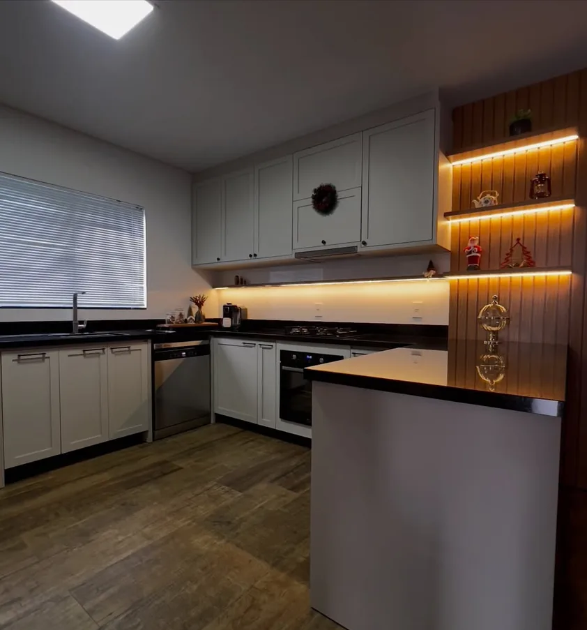 Cozinha em U com armários brancos estilo provençal, bancada escura e prateleiras iluminadas por LED sobre painel ripado de madeira